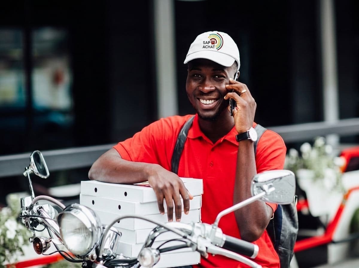 Livreur partenaire en casquette SAP ACHAT, téléphone à la main, colis sur sa moto